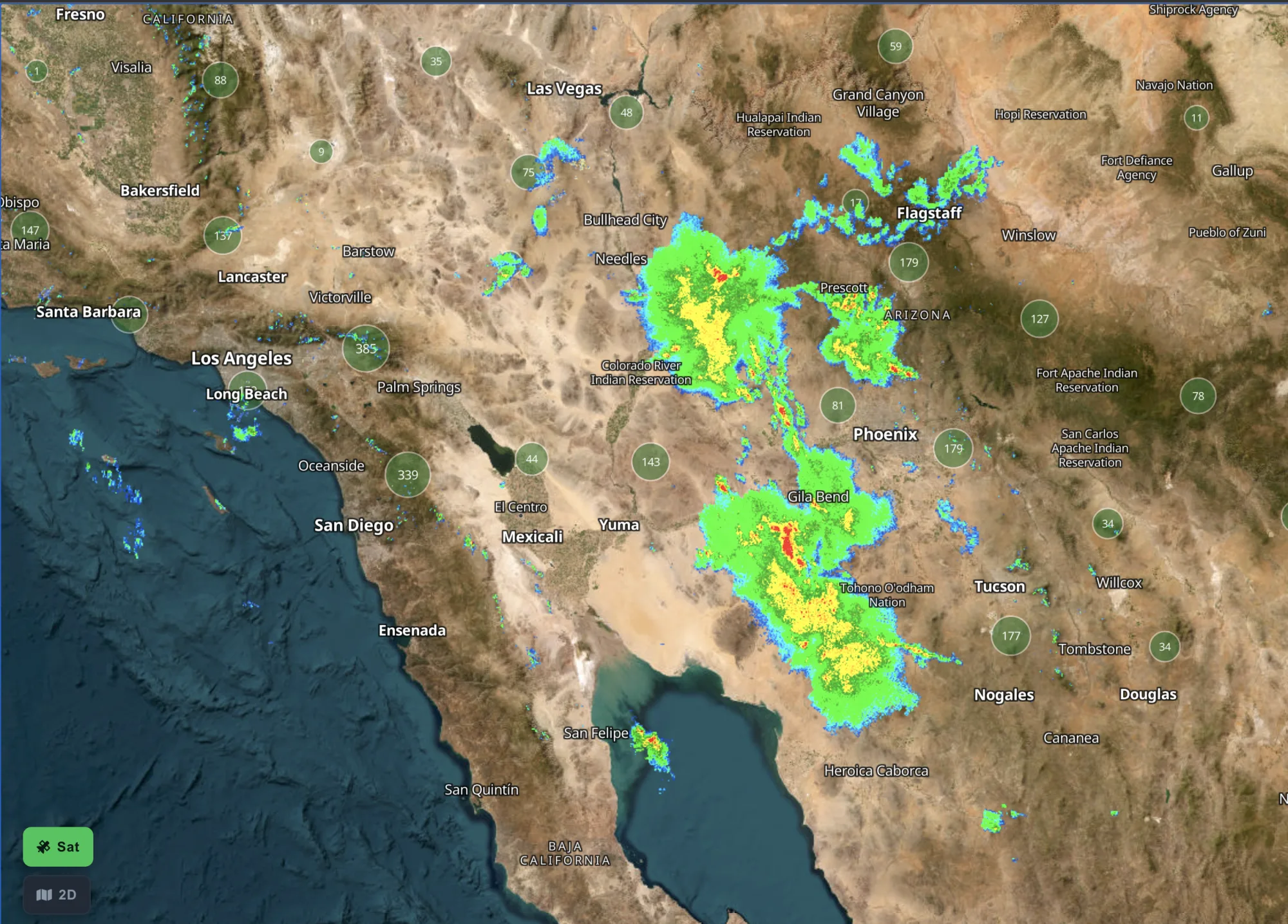 Live rain radar over Arizona and southern California in Motocamp USA: monsoon cells in green, yellow and red spread from Phoenix to Flagstaff and down past Gila Bend, drawn over the satellite basemap with town names still readable on top.